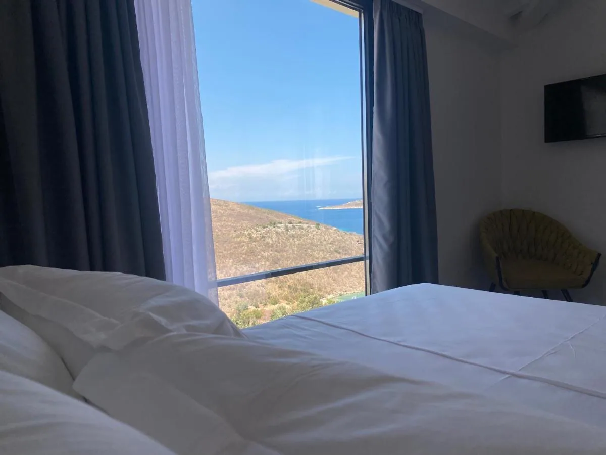 A made bed facing a full-height window onto the hillside and the sea