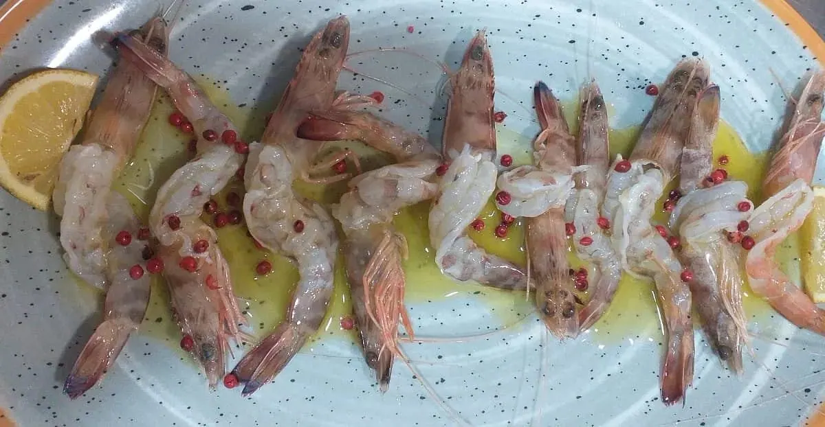A row of prawns scattered with pink peppercorns in olive oil on a pale speckled oval plate, a slice of lemon at one end