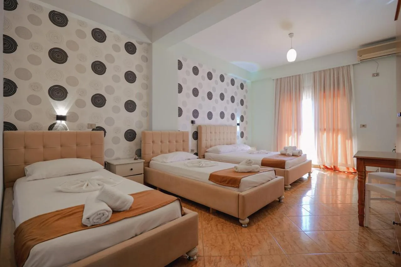 A large bedroom in the original finish: three beds with panel-stitched tan headboards, circle-motif wallpaper, a terracotta-coloured tiled floor and a curtained balcony door.