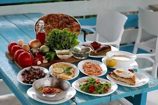 A blue table set with a round flatbread, whole tomatoes, onions, garlic, peppers and a lettuce on a board, a bottle of olive oil, and small plates of olives, salad, yoghurt and a fried egg