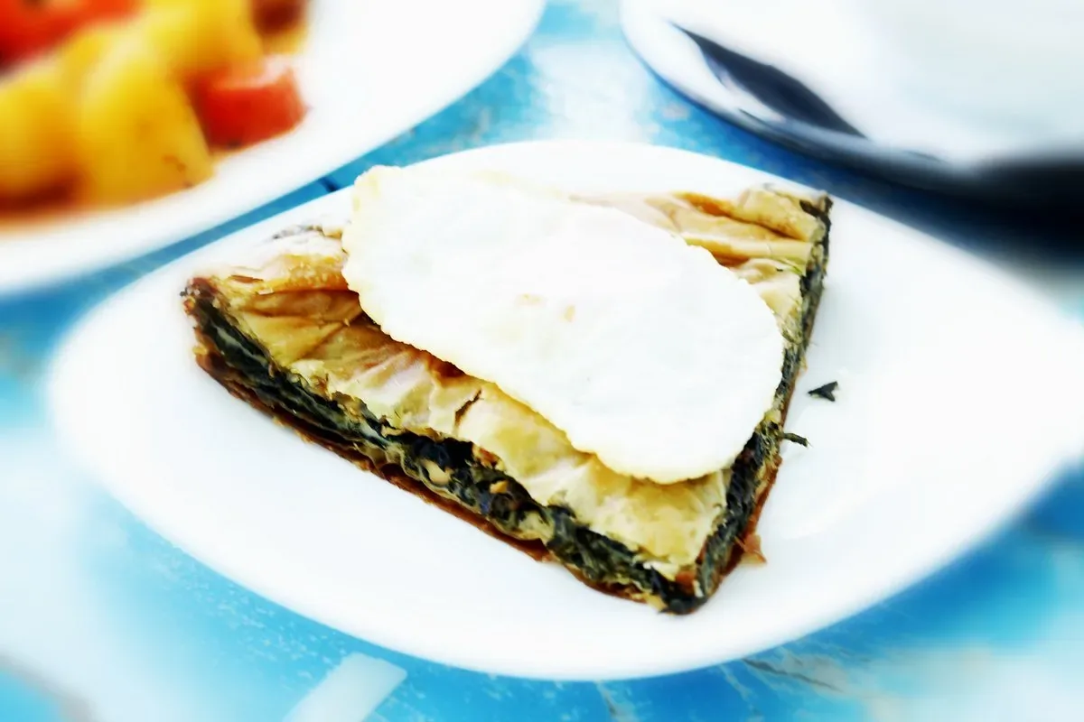 A wedge of dark greens pie in thin filo pastry on a white plate, topped with a fried egg