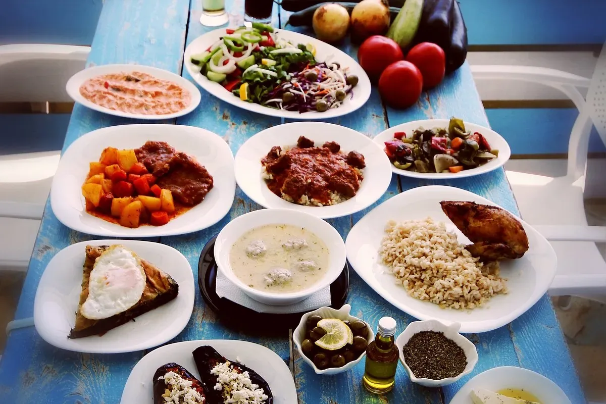 A blue-painted plank table covered with white plates: a chopped salad, a meat stew with potatoes, stuffed peppers, meatball soup, roast chicken with rice, a wedge of pie under a fried egg, stuffed aubergines, olives, olive oil and dried oregano