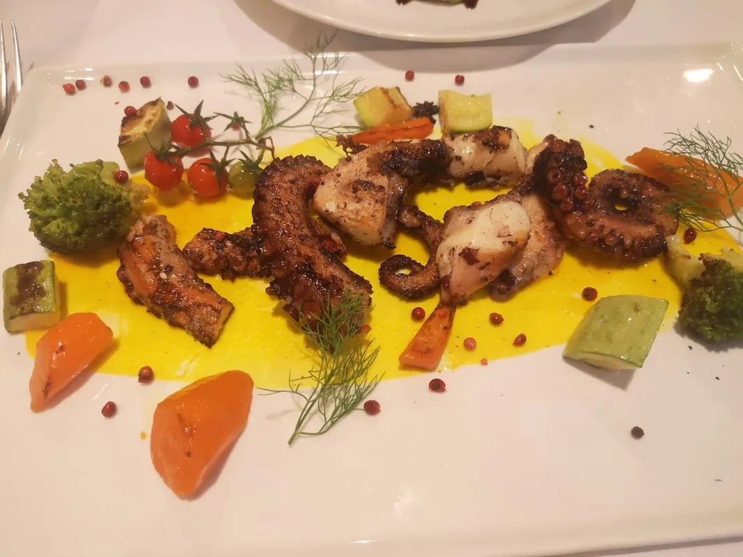Grilled octopus on a saffron-yellow purée with courgette, carrot, broccoli, cherry tomatoes on the vine, pink peppercorns and feathery green fronds on a long white plate