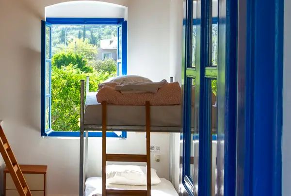 A bunk bed beside an open blue-framed window at Himara Hostel, with garden greenery outside