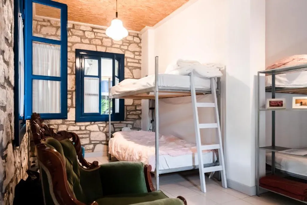 Dormitory room at Himara Hostel with metal bunk beds against a bare stone wall, blue-framed windows and green velvet armchairs
