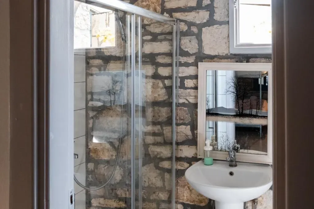 Shower room at Himara Hostel with a glass screen against an exposed stone wall and a basin under a framed mirror