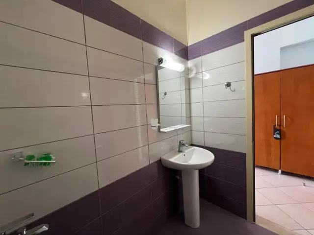 A bathroom at Hotel Prinos tiled white above and aubergine from dado height down to the floor, with a second aubergine course under the ceiling, a pedestal basin, a mirror and a wall light
