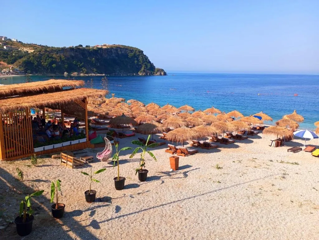 A shingle beach with rows of straw parasols and sun loungers, a timber beach bar, and a green headland closing the bay