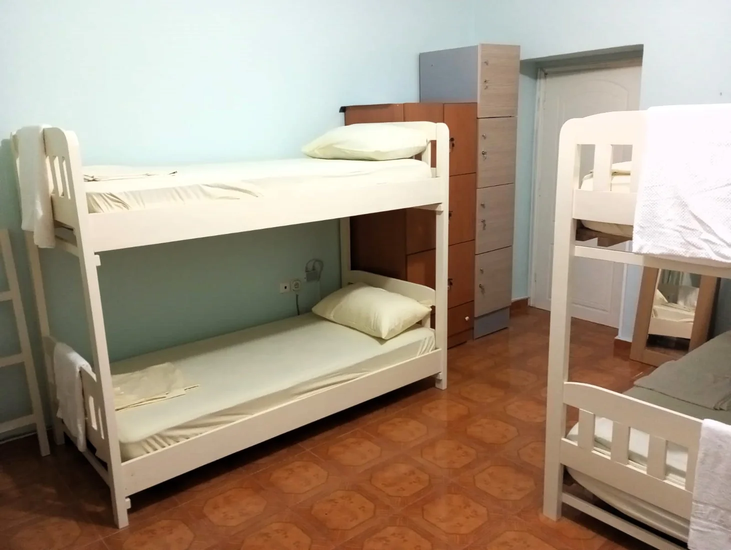 A mixed dormitory with three pine bunk beds, a tall wooden locker column and a tiled floor