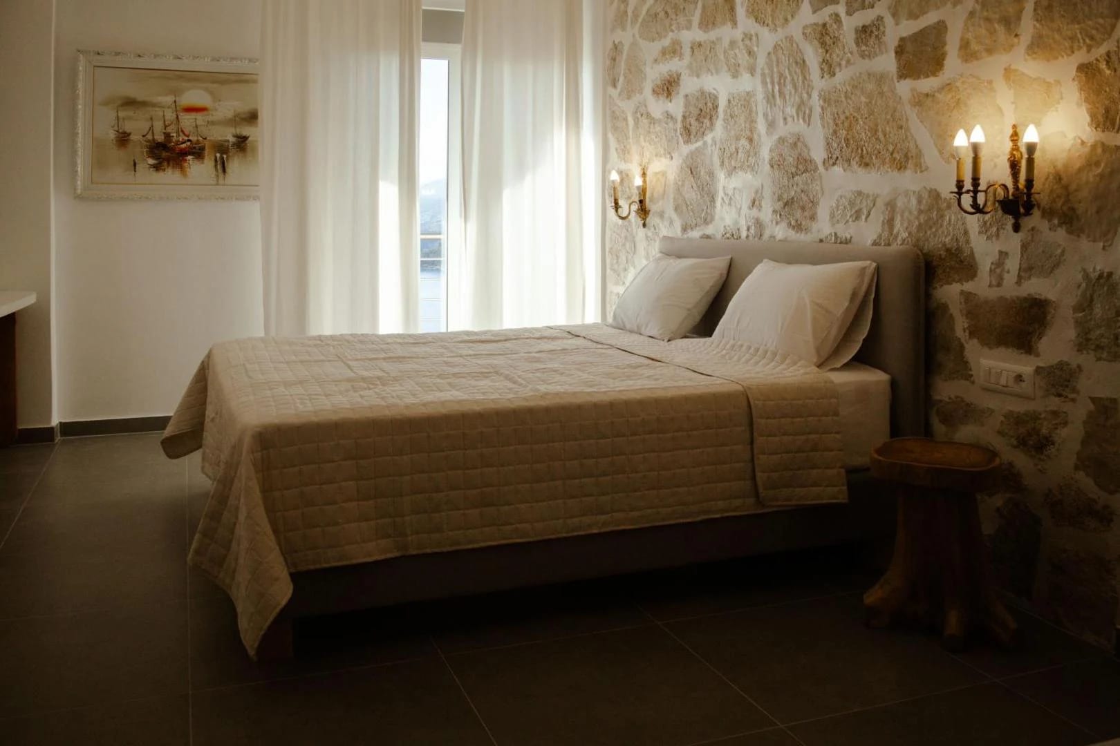 A double bed with a linen headboard against a stone wall, a window beside it looking out to the sea