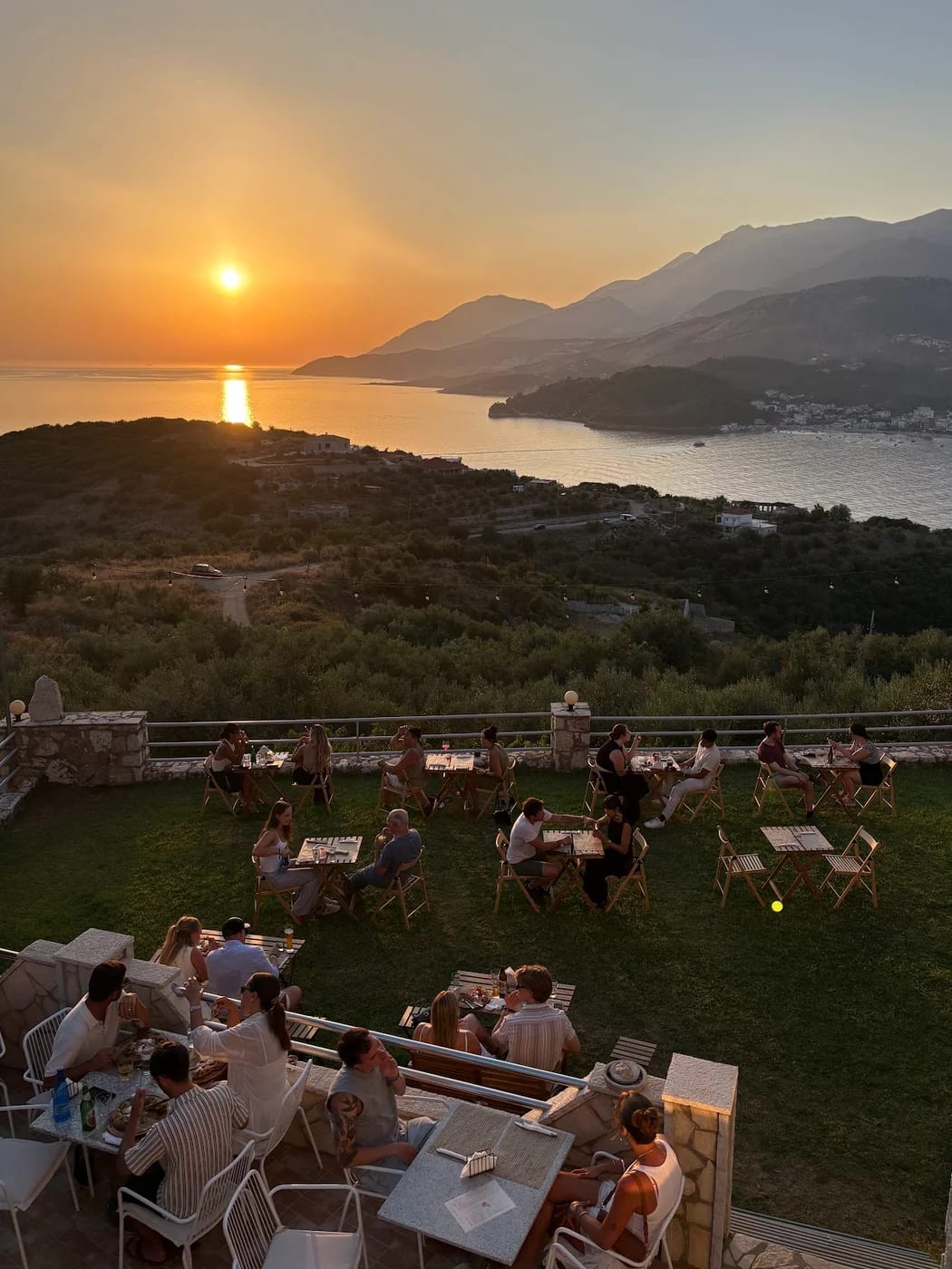 Wooden tables on a grass terrace above Himarë bay as the sun drops to the sea