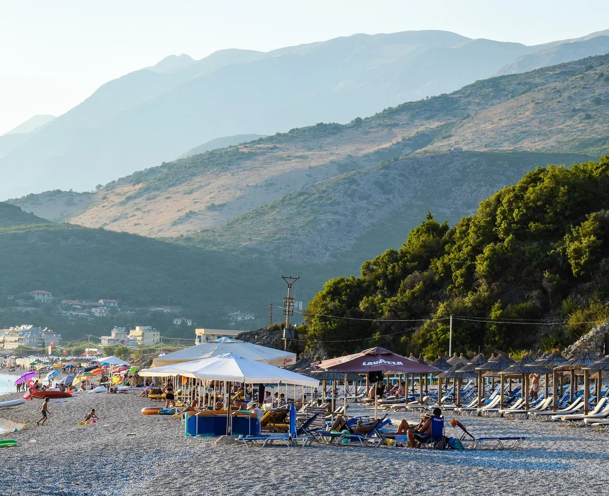 Livadhi Beach in late afternoon: grey shingle, straw and canvas parasols, sun loungers, and the mountains behind Himarë.