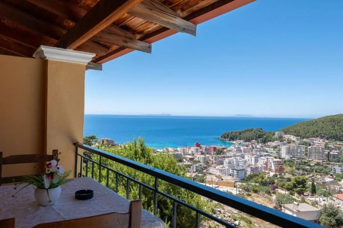 A balcony table under a timber canopy with Himarë town, the bay and the headland spread out below