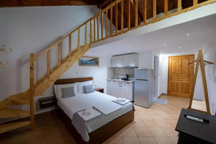 A double bed beside a pine staircase climbing to a mezzanine, with a kitchenette and fridge along the far wall