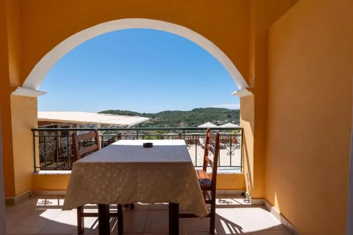 A table and two chairs set inside an arched balcony with an ochre wall, the pool deck and the wooded hills beyond