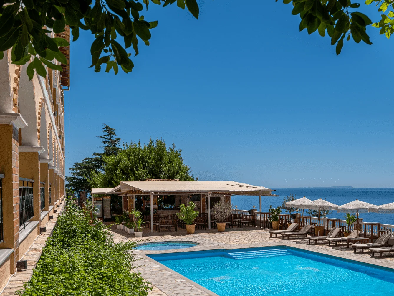 The pool deck framed by overhanging leaves, the arcade and its wrought-iron rails on the left, open sea at the end