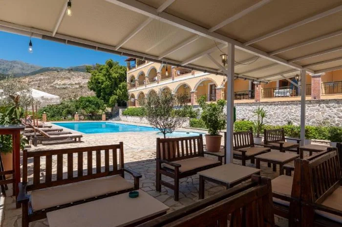 Wooden benches with cream cushions under the pool bar's canopy, looking across the water to the arcade and the ridge behind