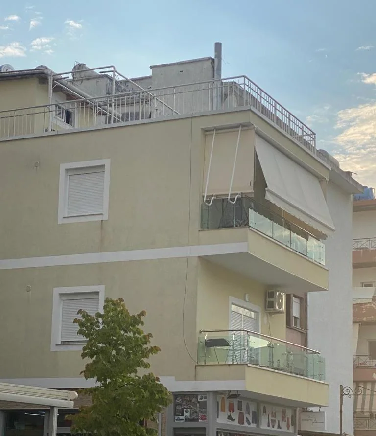 The cream-yellow apartment block at dusk, with glass-balustraded balconies, an awning over the top one and shops at street level