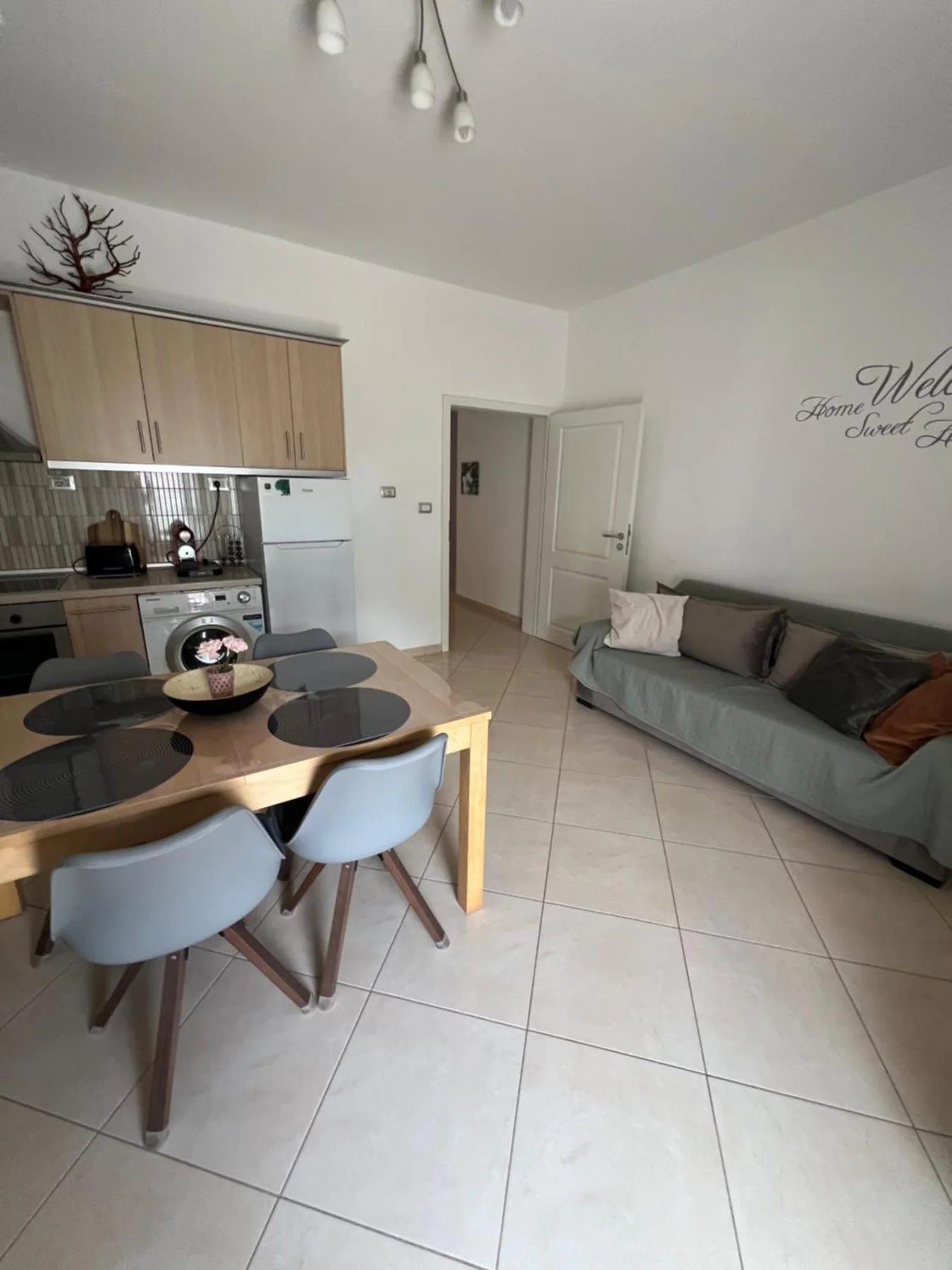 The living room with a grey sofa against the wall, tiled floor, and an open doorway through to the hall