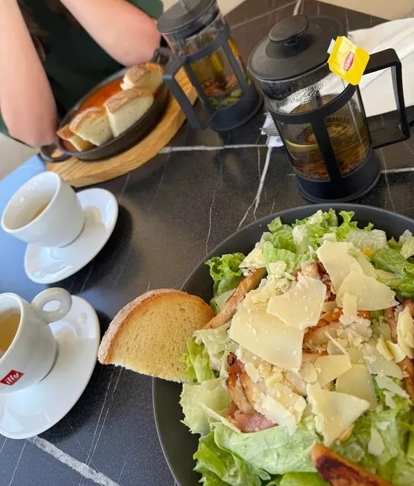 A caesar salad with shaved parmesan on a dark marble table, beside two glass tea presses and espresso cups