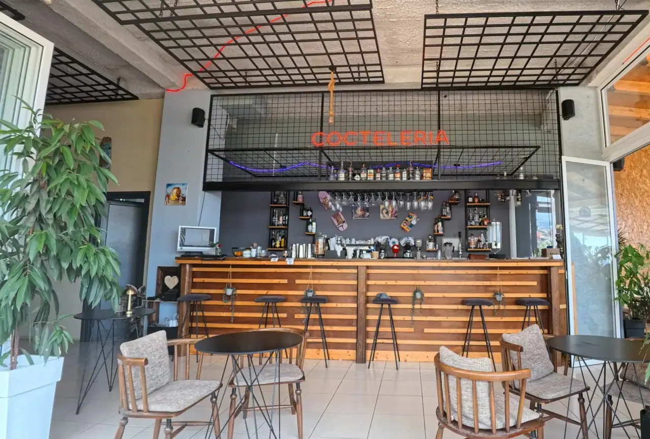 The bar counter in timber slats under a black steel mesh ceiling, with a red neon Cocteleria sign and a violet neon tube