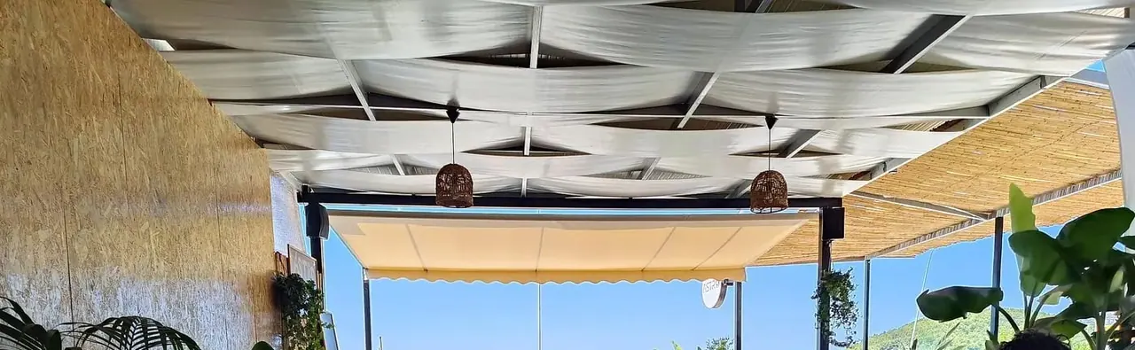 White sailcloth strung in a lattice over the terrace, with rattan pendant lamps, a cream awning and a wooded hillside beyond