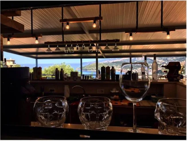 Glasses on a bar counter under a boarded timber roof, stemware hanging from a rail above, and Himarë Bay beyond the open side