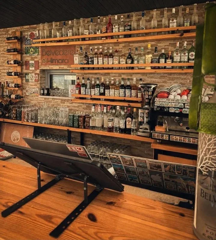 The bar: pine shelves of bottles against a split-stone wall, novelty licence plates for Tokyo, Brazil, Peru, Mexico and Medellín on the left, and the hand-painted Belvedere Himara board with its tree roundel between two of the shelves