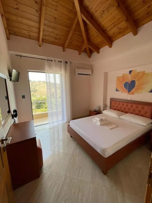 A double bed with a rust buttoned headboard under exposed pine rafters, a desk and mirror to the left and a curtained balcony door onto the green hillside