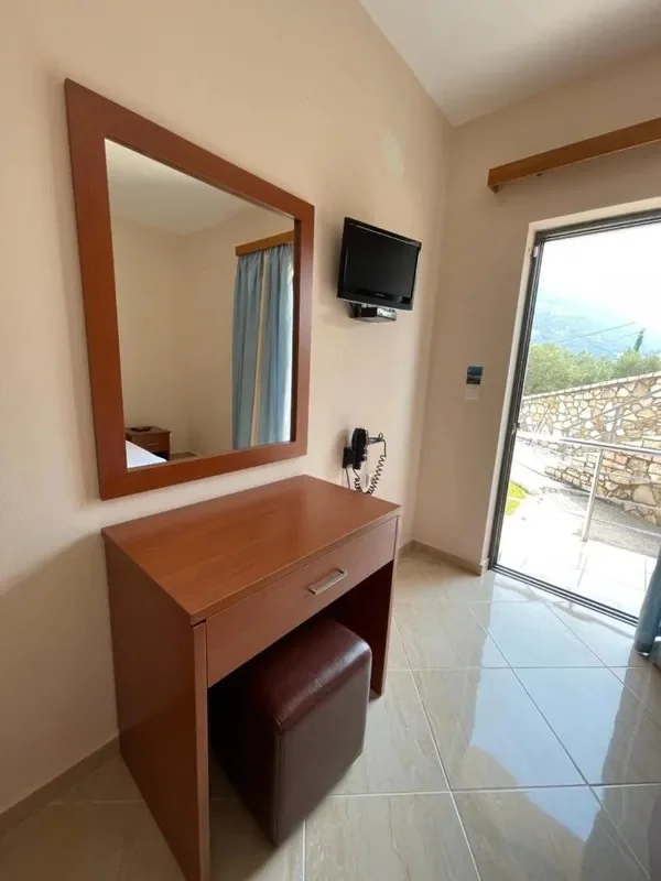 A desk and mirror beside a flat-screen television and a sliding balcony door opening onto a stone wall and the slope beyond