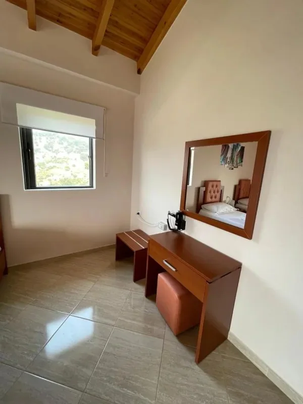 A wooden desk, stool and framed mirror against a plain wall, under exposed pine rafters, with a small window onto the hillside