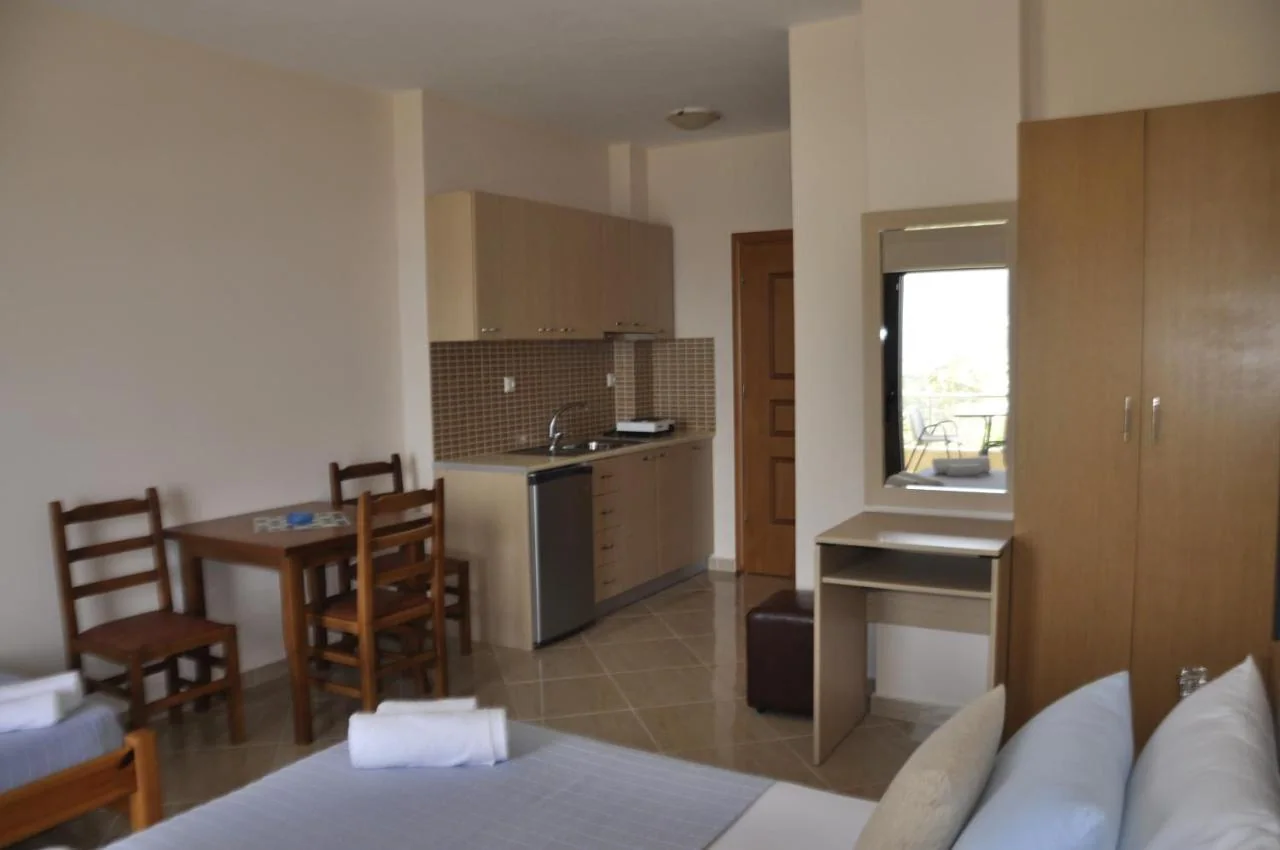 An apartment interior with a fitted kitchenette, a fridge, a wooden dining table and four chairs, and a window through to the balcony