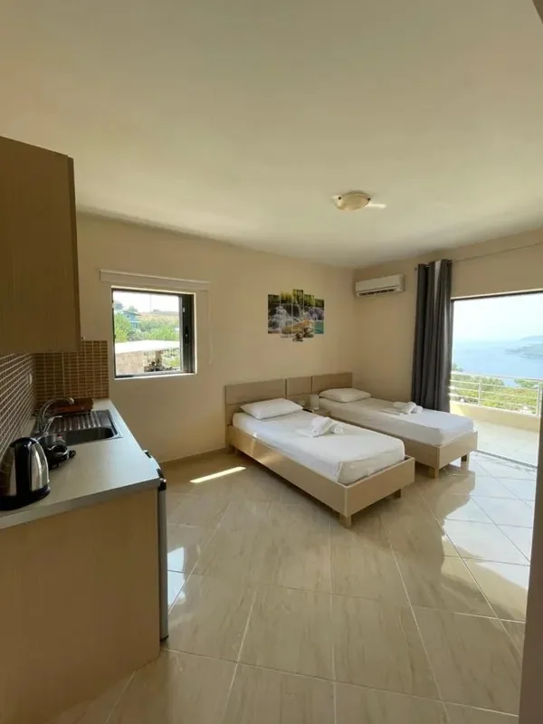Two single beds side by side in a tiled room with a kitchenette and kettle on the left and the sea visible through the balcony door