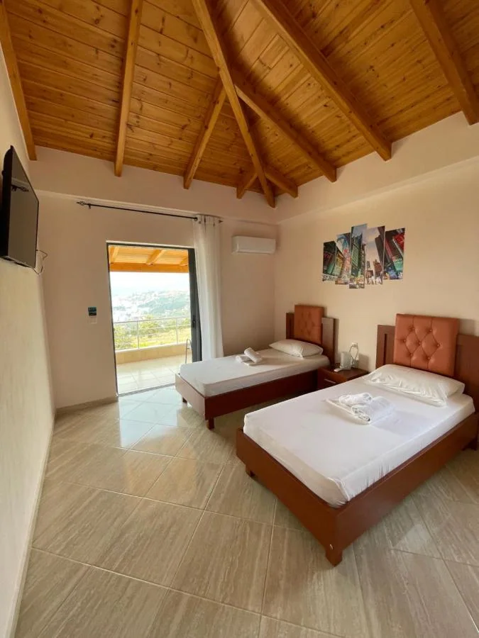 Two beds with rust-coloured buttoned headboards under a full pine A-frame roof, an air-conditioning unit on the wall and a balcony door open onto the town and the bay