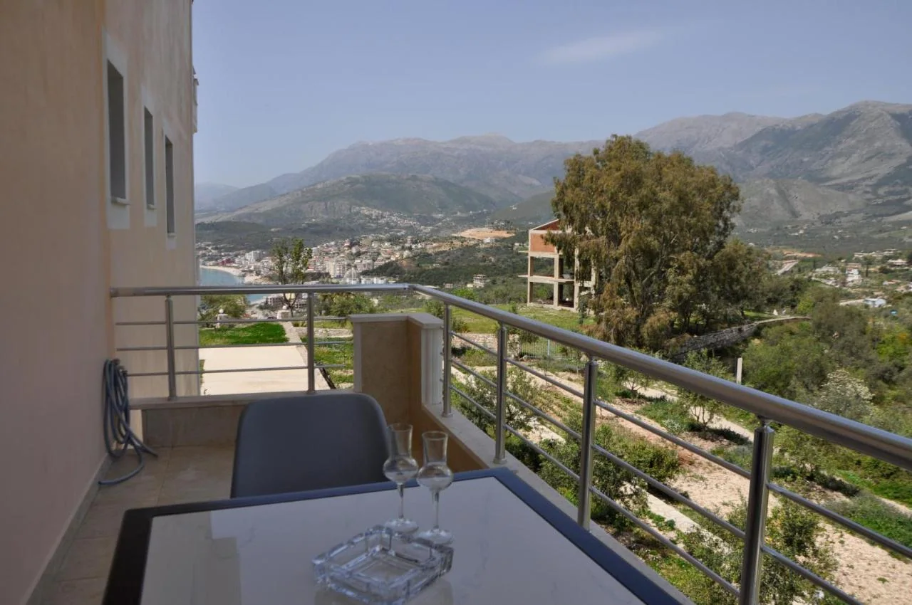 A balcony table with two glasses and an ashtray in the morning, Himarë and its beach on the left and the bare mountain ridge across the valley
