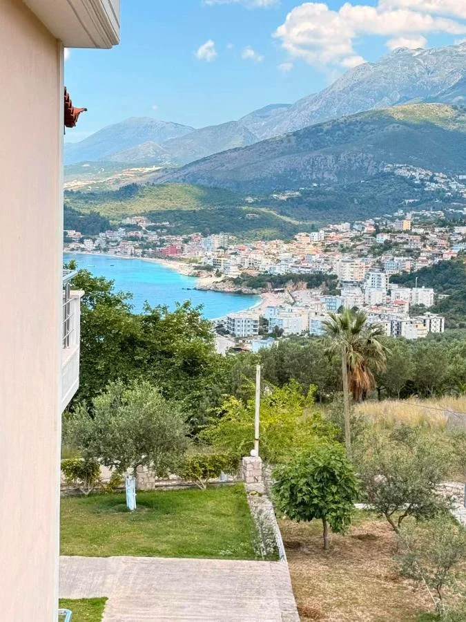 Himarë town and its turquoise bay seen over the property's garden from the floor above it, with the mountain wall rising behind the town