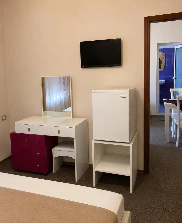 A dressing table with mirror and a magenta chest of drawers, a fridge and wall-mounted television, with a blue-tiled bathroom through the doorway.