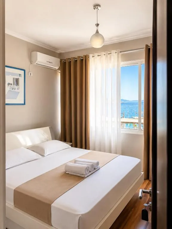 A double bed made up in white and beige beside a curtained window showing the sea, with a wall air-conditioning unit high on the side wall.