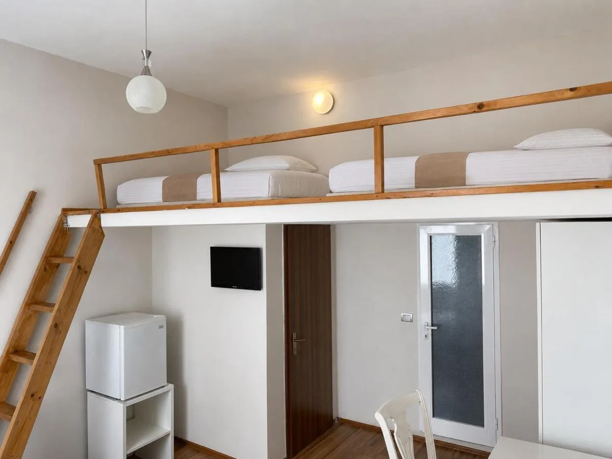 A room with a timber ladder rising to a railed mezzanine platform carrying beds made up in white, a television and a fridge below.