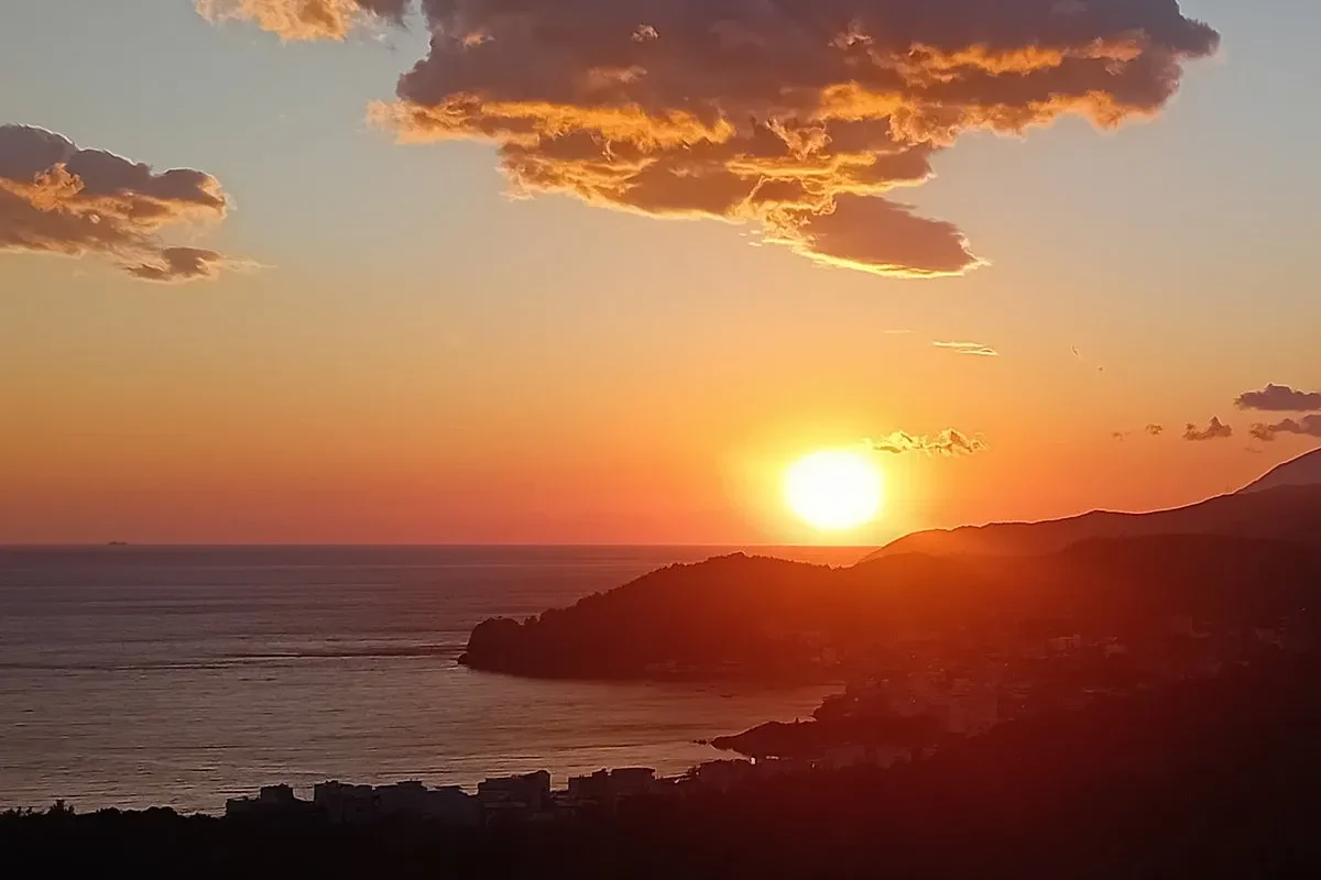 Sunset over the Ionian: the sun dropping onto the ridge of a dark headland, open water to the left of it, and a town strung along the shore below