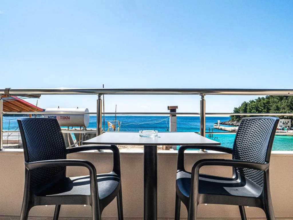 Two chairs and a table on a balcony with the sea and a small jetty beyond the railing
