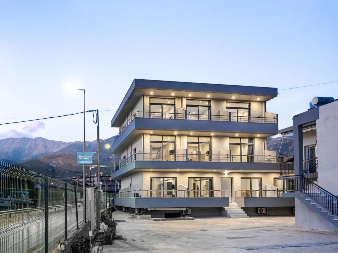Endless Blue Himara at dusk, three storeys of lit balconies above its own parking forecourt with the mountains behind
