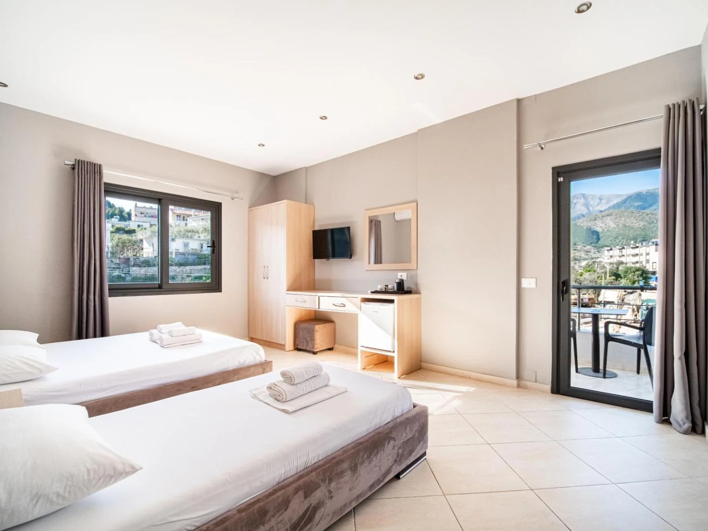 A room with two single beds, a desk and a balcony looking towards the mountains behind Himarë