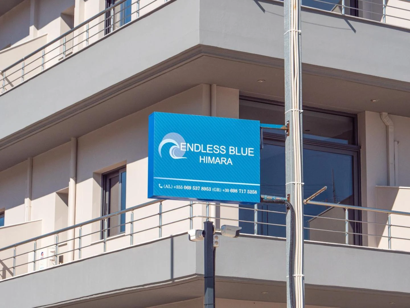 The Endless Blue Himara sign, white lettering and a wave mark on a blue panel, bolted to a pole in front of the balconies