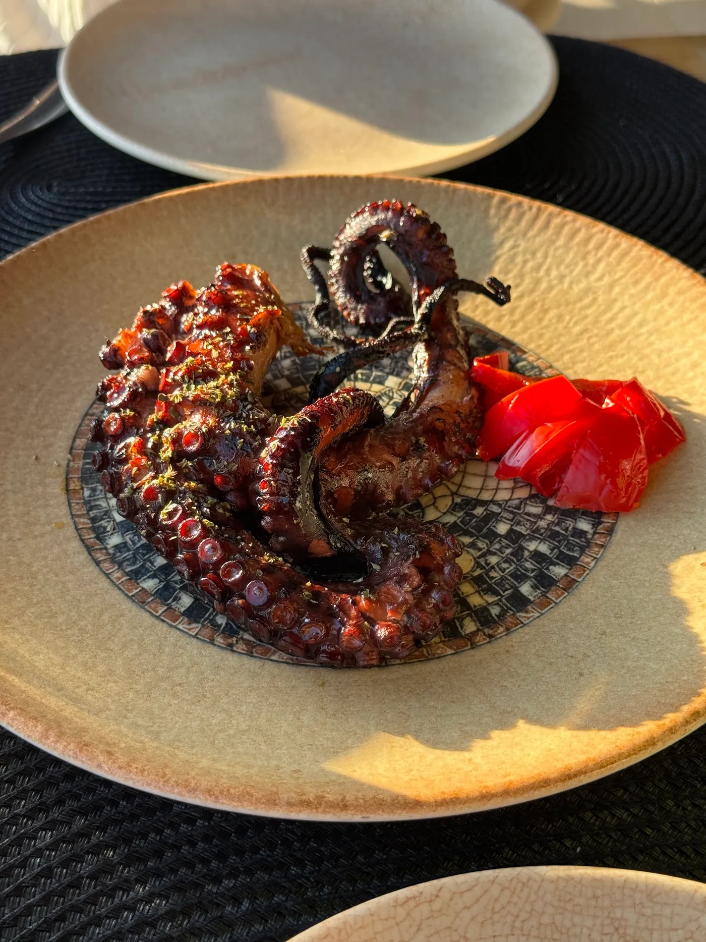 Charred grilled octopus with peeled roast red peppers, on a sand plate with a blue-and-white printed centre, at Fig & Olive, Himarë