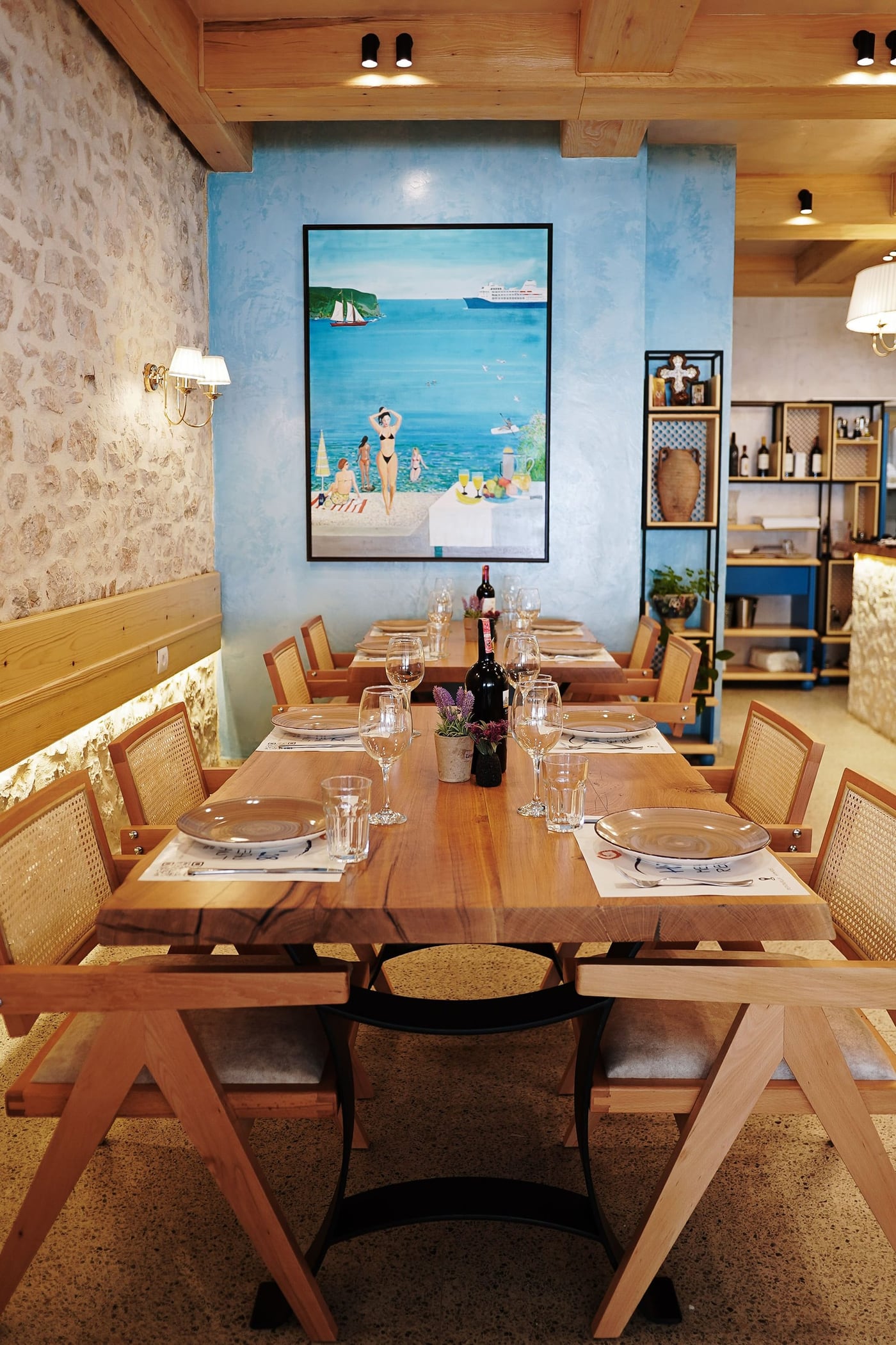 Dining room with a stone wall, pale blue plaster, timber ceiling beams, live-edge tables and cane chairs, a painted beach scene on the wall