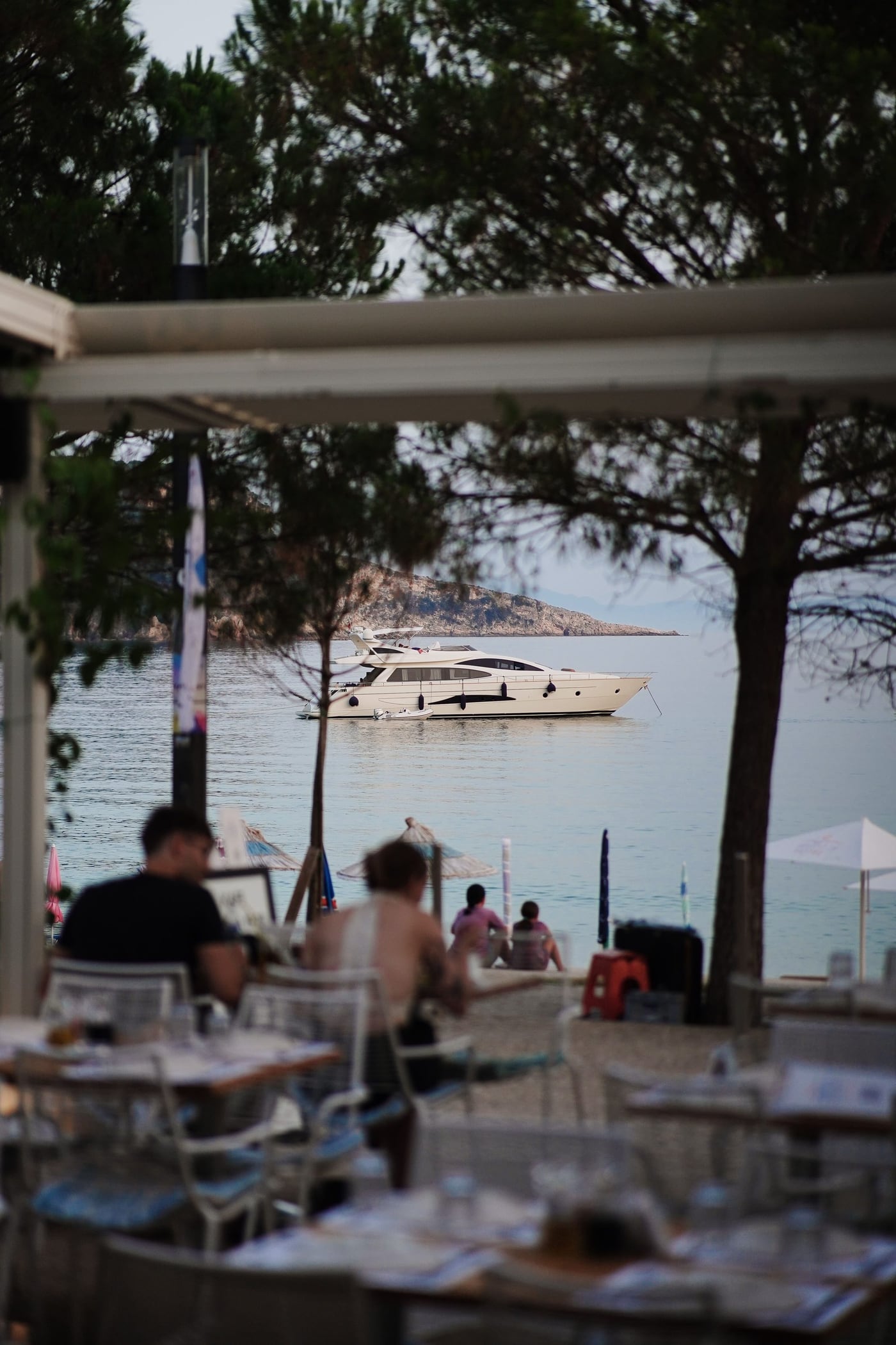 Looking out over the terrace tables and the beach beyond them to a moored motor yacht on flat water, a rocky headland behind and pines framing the view
