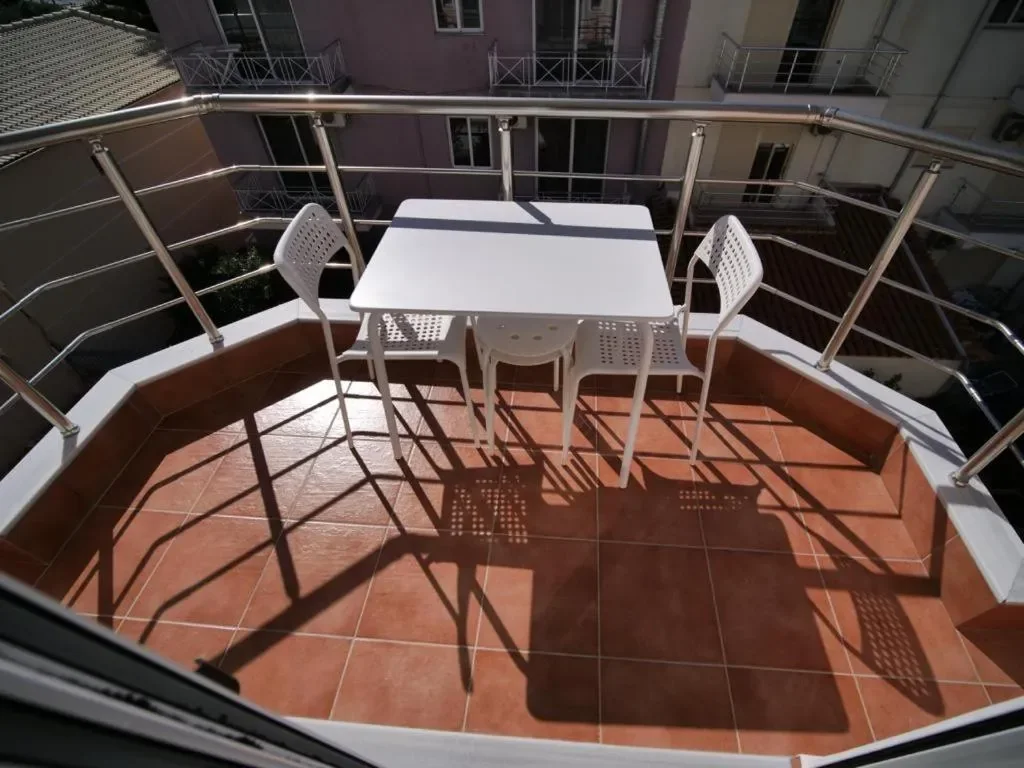 A faceted hotel balcony seen from above, terracotta tiles, a white table with two chairs and a stool, and a steel rail