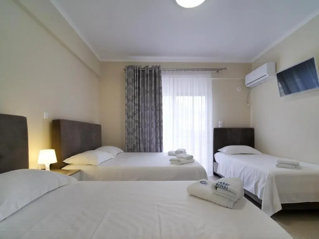 Three beds in a cream-walled room, a crown and the word ROYAL embroidered on the folded towels nearest the camera, a floor-length curtain behind