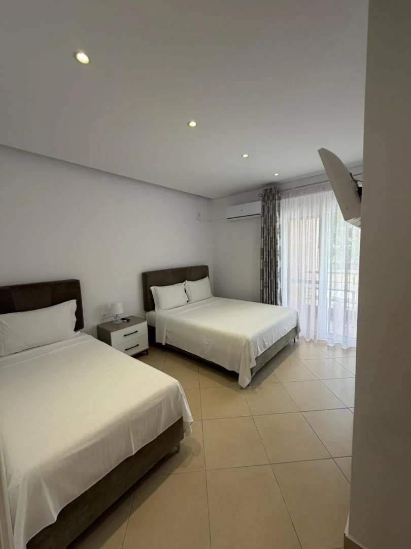Two large beds in a white room with a wall air-conditioning unit and a curtained balcony door